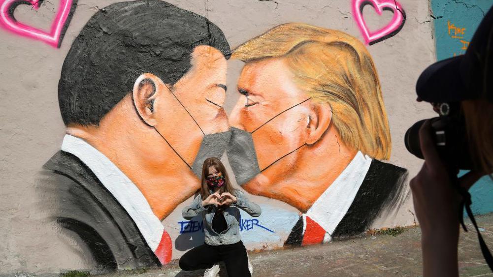 A young woman named Lily wears a face mask as she poses for her friend in front of graffiti of Communist Party of China General Secretary Xi Jinping (L) and U.S. President Donald Trump kissing each other while wearing face masks during the coronavirus crisis on April 27, 2020 in Berlin, Germany.