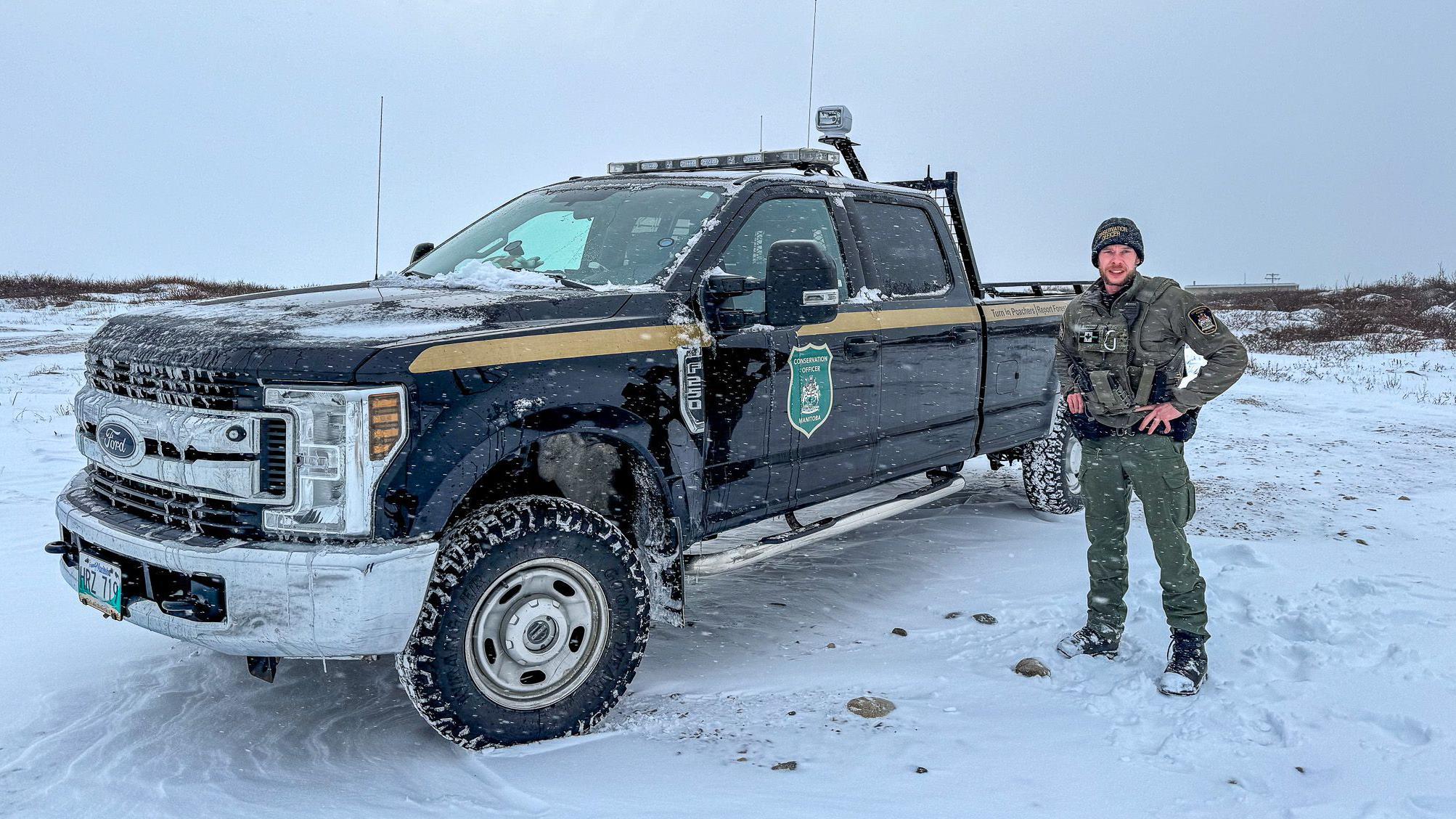 A imagem mostra o sargento Ian Van Nest, do Programa de Alerta de Ursos Polares de Churchill, parado ao lado de uma caminhonete preta oficial em um ambiente nevado, equipada para patrulha e segurança em áreas onde os ursos polares estão presentes.