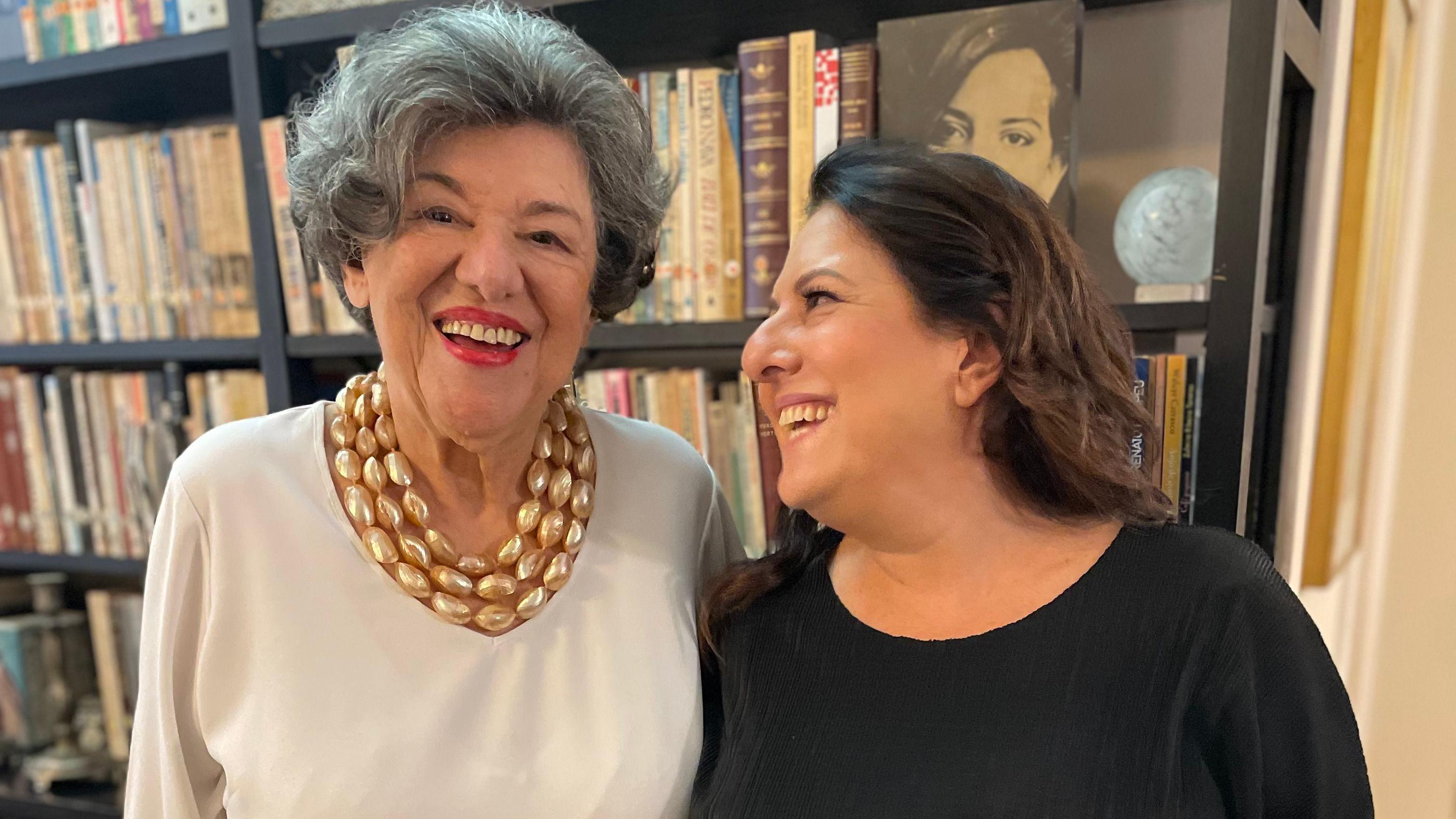 Ruth e Mariana abraçadas e sorrindo em frente a estante de livros