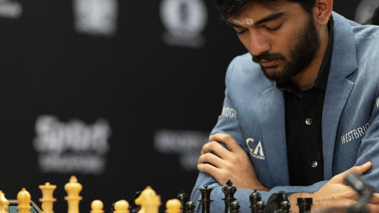 Gukesh Dommaraju mira el tablero de ajedrez durante la partida del jueves. Lleva una chaqueta azul y una camisa negra y tiene los brazos cruzados, con los codos apoyados sobre la mesa.