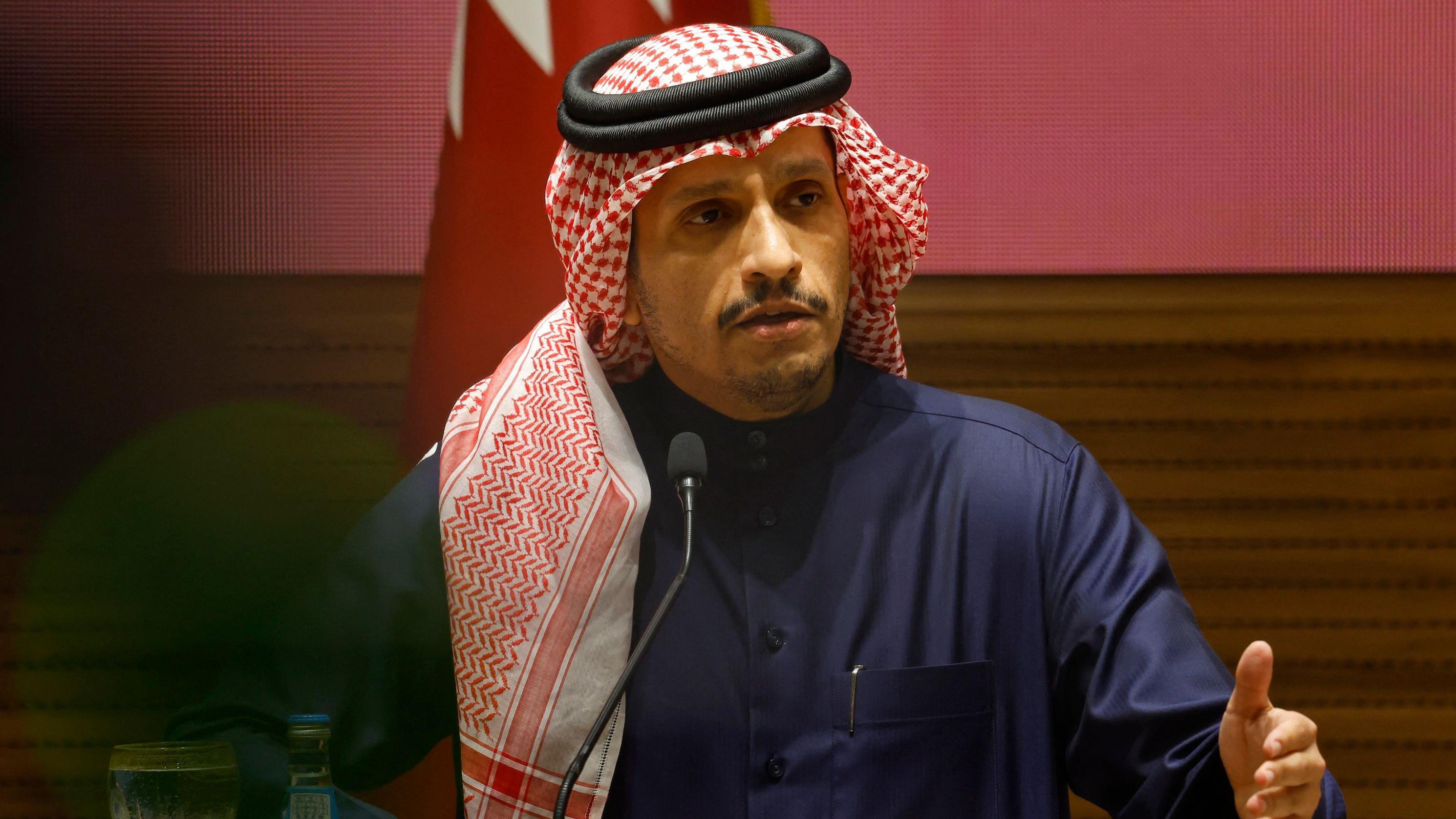 Mohammed bin Abdulrahman bin Jassim al-Thani gesticulando enquanto fala ao microfone, em frente à bandeira 