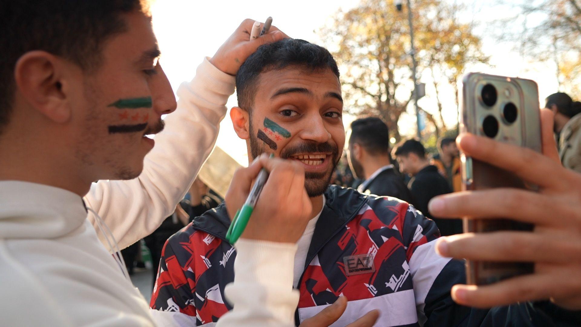 Un hombre pinta la bandera verde, blanca y negra de la antigua República Siria en la cara de otro hombre.