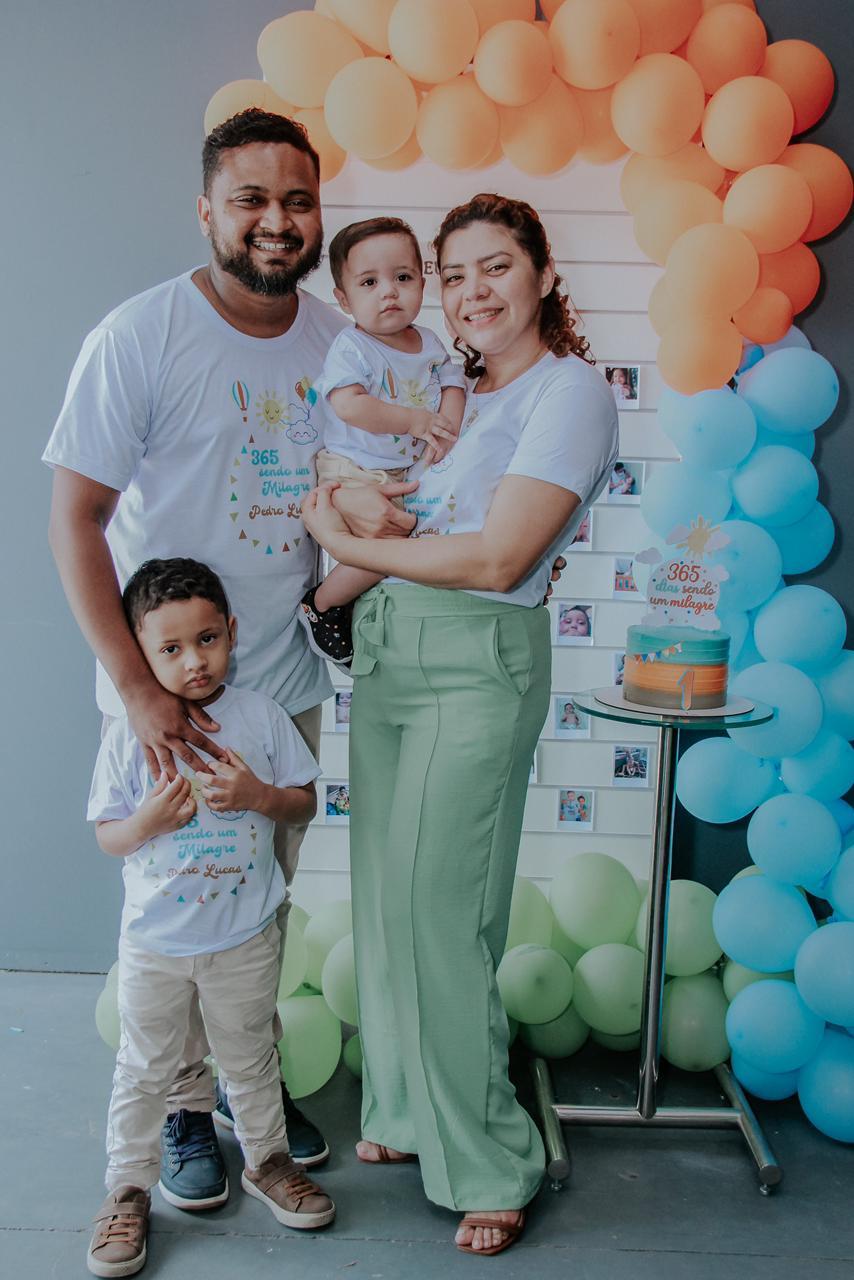 Pedro no colo da família em frente a mesa com bolo de aniversário e balões coloridos