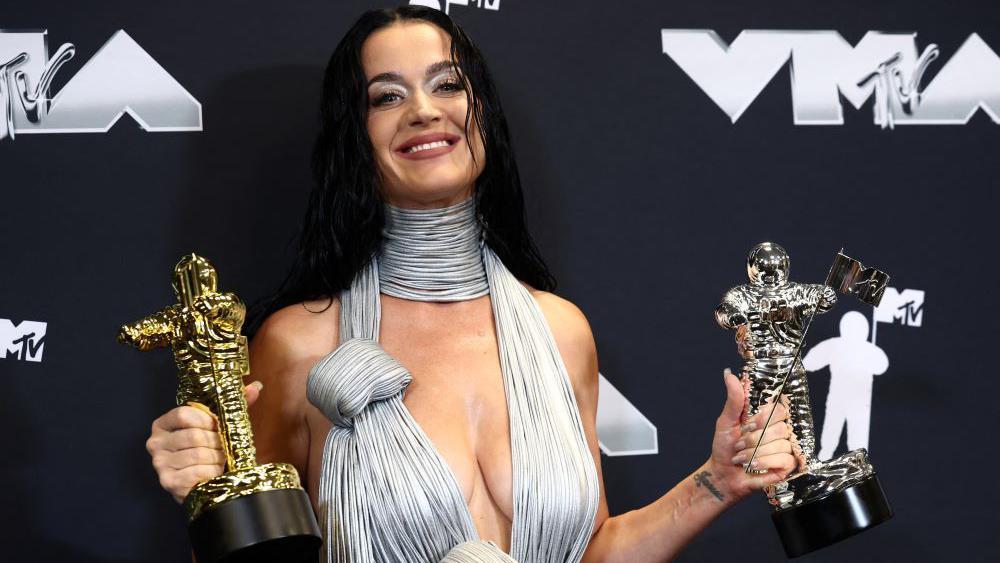 A foto mostra a cantora Katy Perry da cinctura para cima, vestindo uma roupa prateada decotada e segurando dois troféus, um prateado e um dourado. 