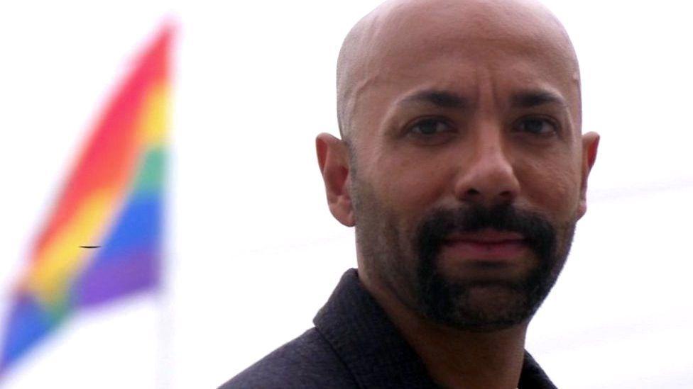 Nas Mohamed olhando com olhar confiante para foto, com bandeira do arco-íris atrás