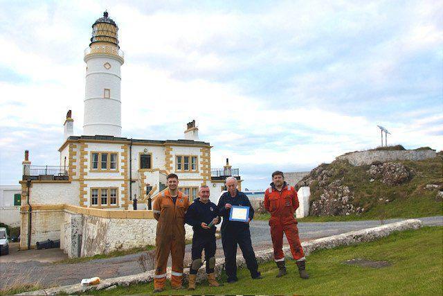 Ross Russell, dois engenheiros e o faroleiro estão do lado de fora do Farol Corsewall. Eles estão vestindo macacões de trabalho. O farol é branco