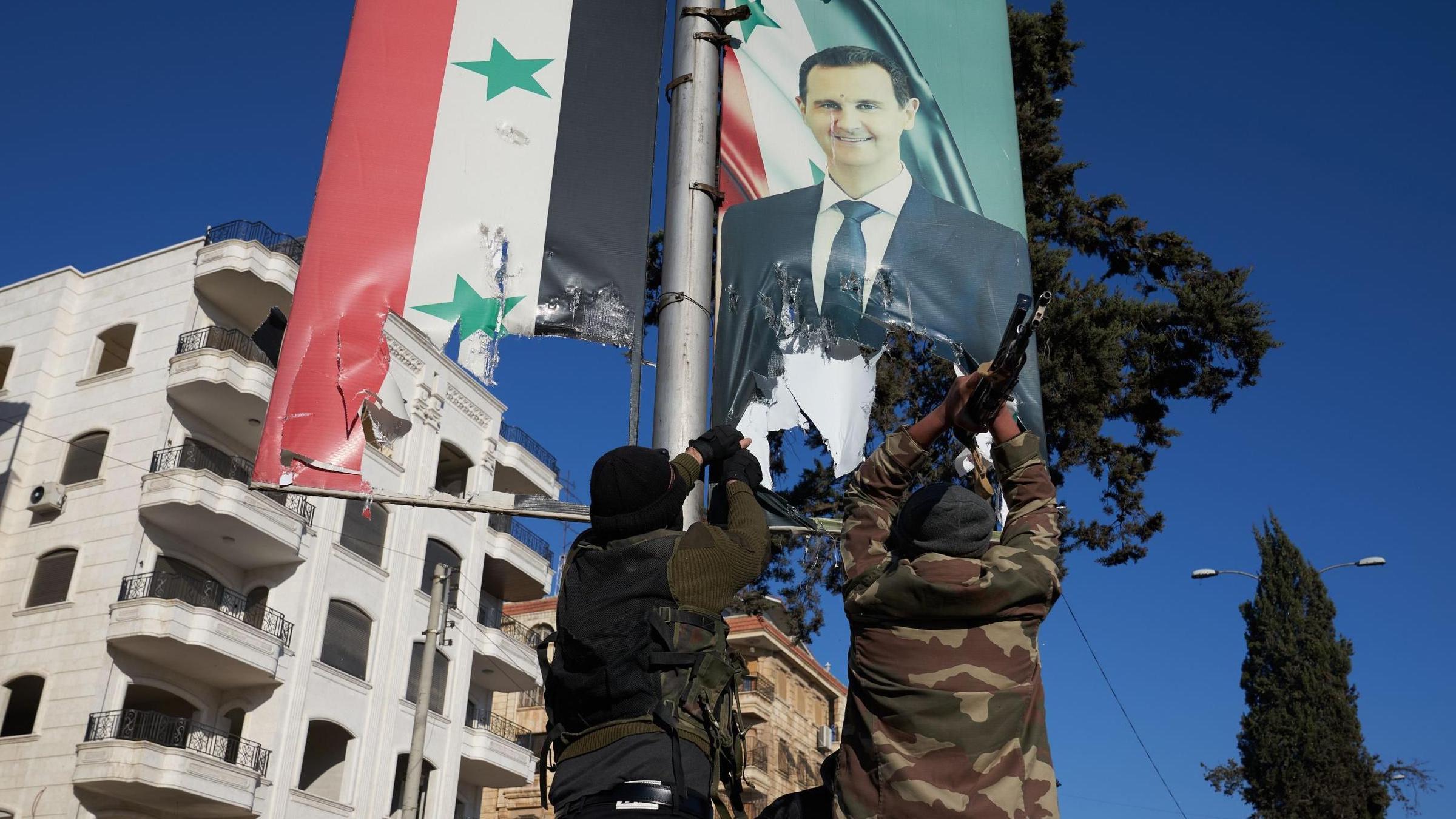 Dos hombres con ropa de camuflaje vistos desde detrás de un cartel con la imagen del presidente sirio, Bashar al Assad.