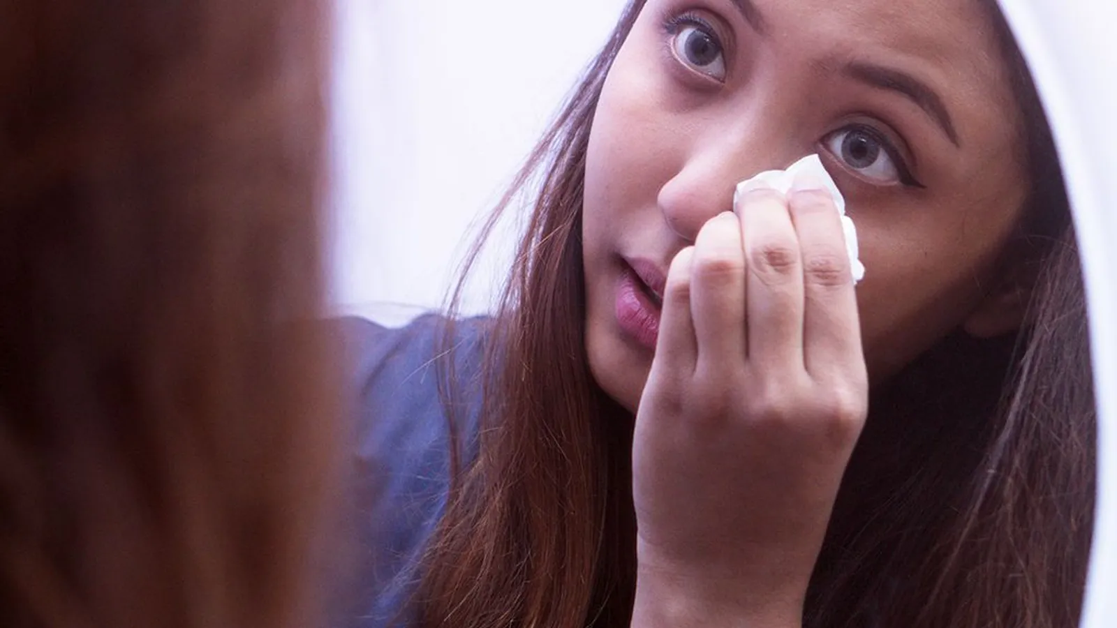 Mulher de feições asiáticas e cabelo castanho-escuro se olha no espelho enquanto aplica um produto com uma gaze