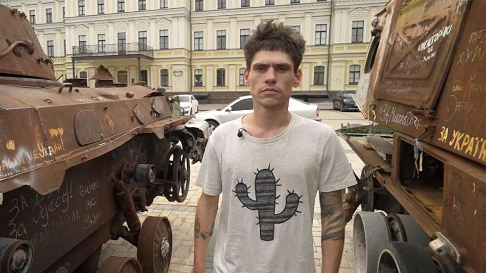 Oleksii de pé entre dois veículos militares em Kiev após seu resgate. Ele tem cabelos castanho curto e veste uma camiseta branca com uma imagem de um cacto