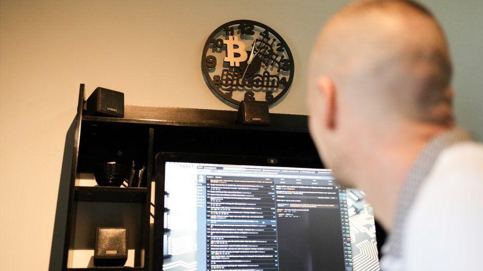 The back of a man's head, looking at a computer screen, with a clock with a Bitcoin logo on the wall 