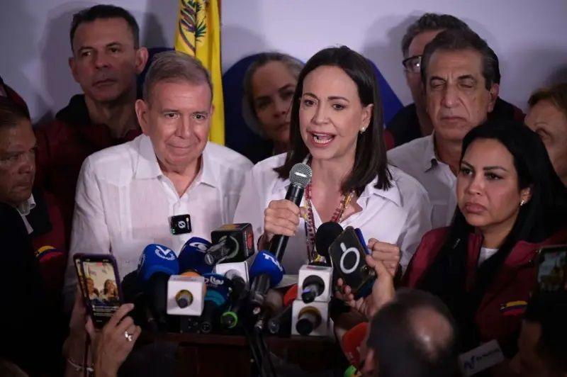 A foto mostra González, um homem de pele clara e com pouco cabelo, e Corina, uma mulher de pele clara e cabelos escuros, ambos de camisa branca. Corina fala, rodeada de microfones. 