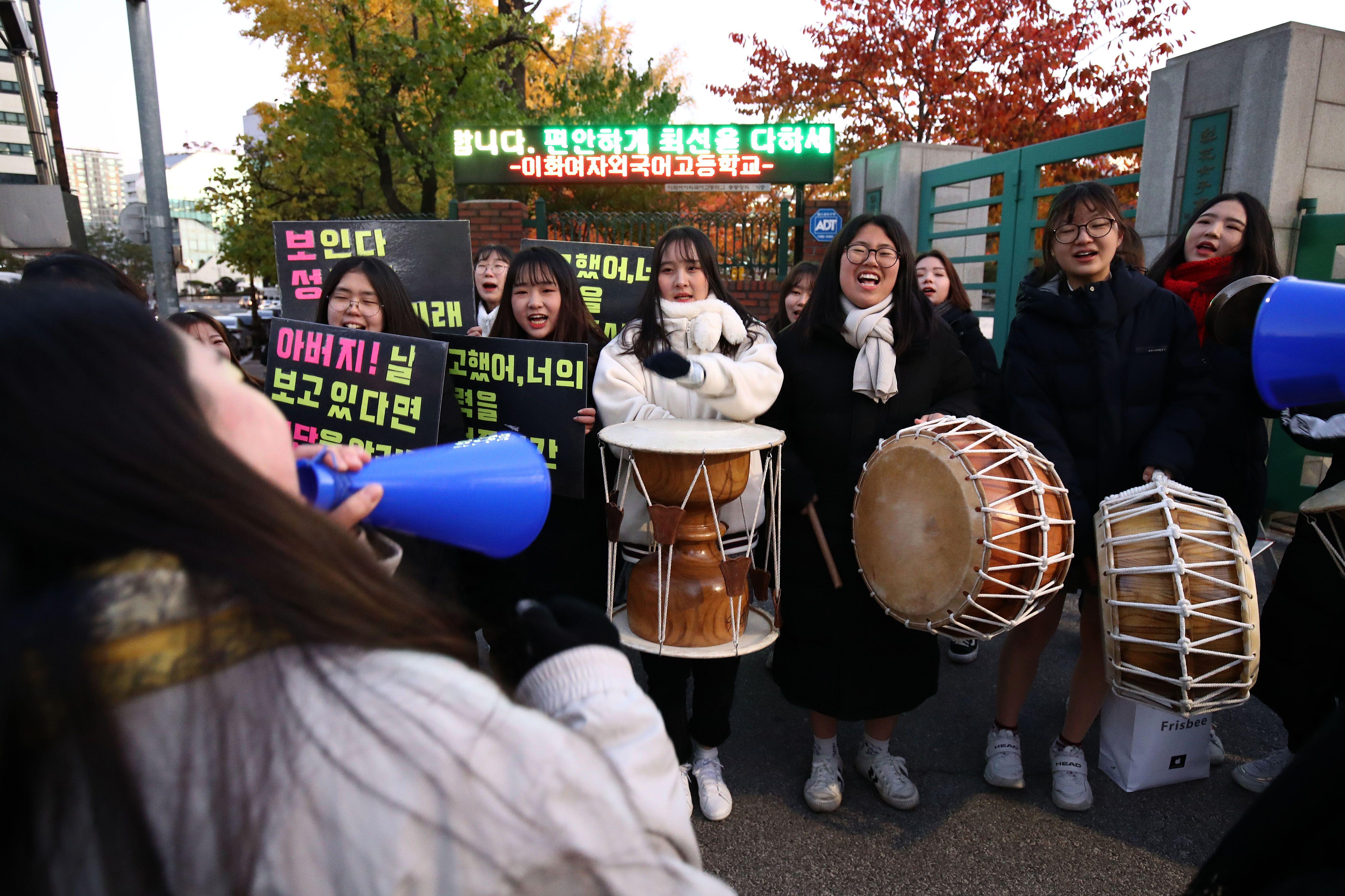 Alunos esperando do lado de fora dos centros de testes com cartazes e instrumentos musicais