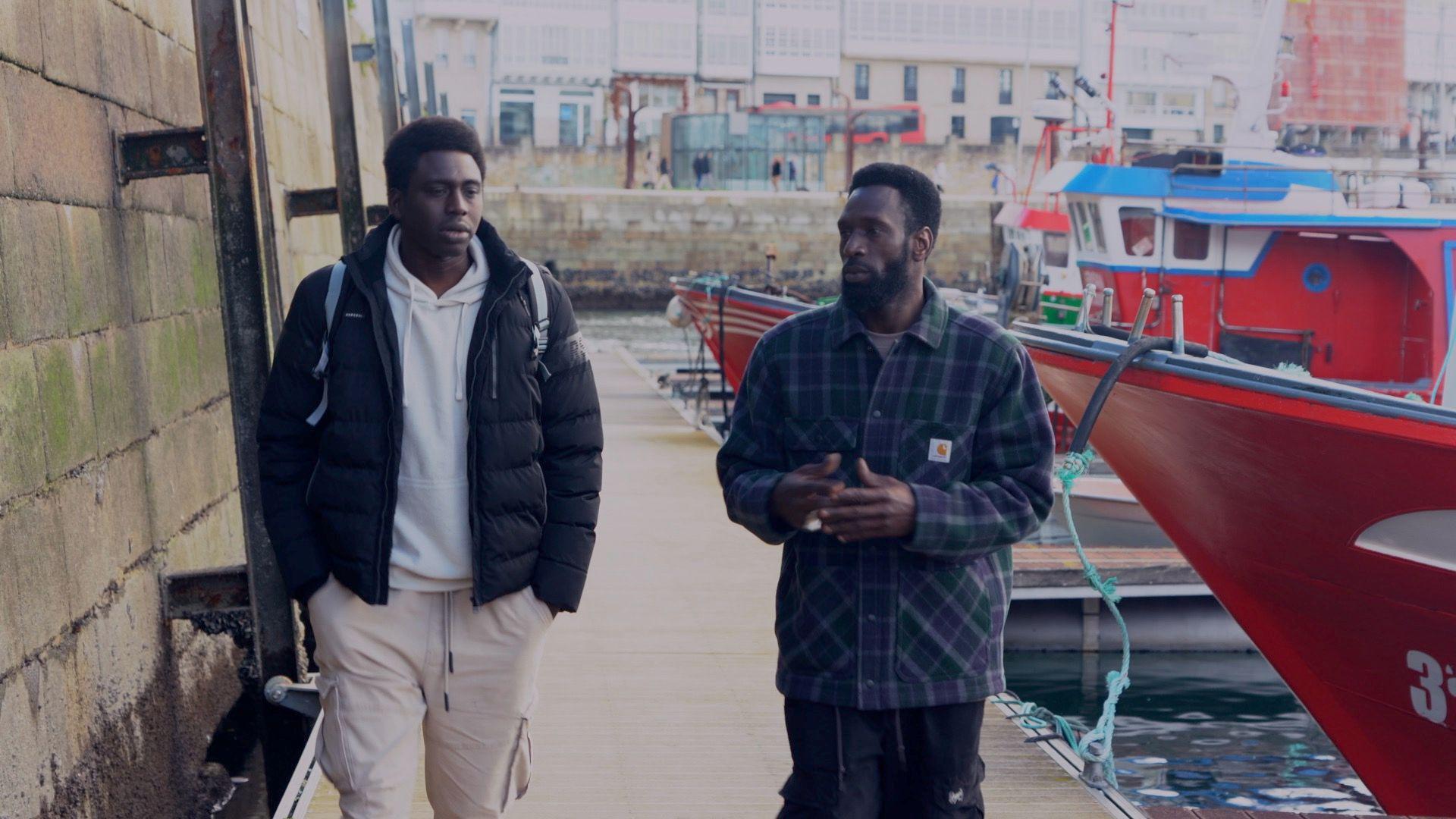 Ibrahima Diack y Magatte Ndiaye charlando enfrente de varios barcos de pesca rojos.