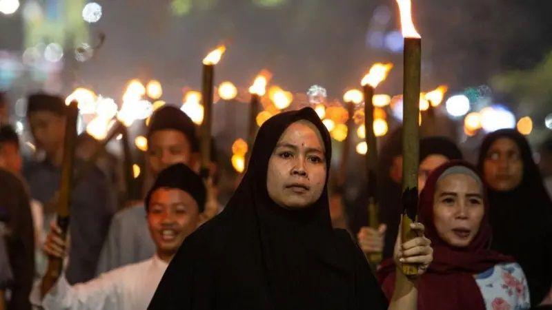 Em primeiro plano, uma mulher com a cabeça coberta, com uma tocha na mão, seguida por diversas pessoas atrás.