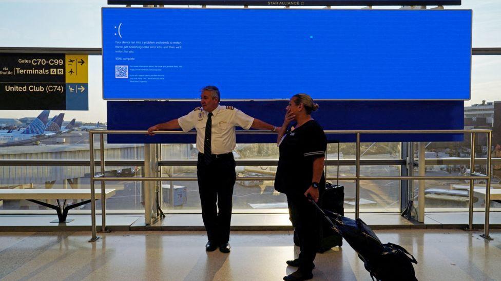 Una pasajera y un funcionario de aerolínea parados frente a una pantalla azul de un aeropuerto