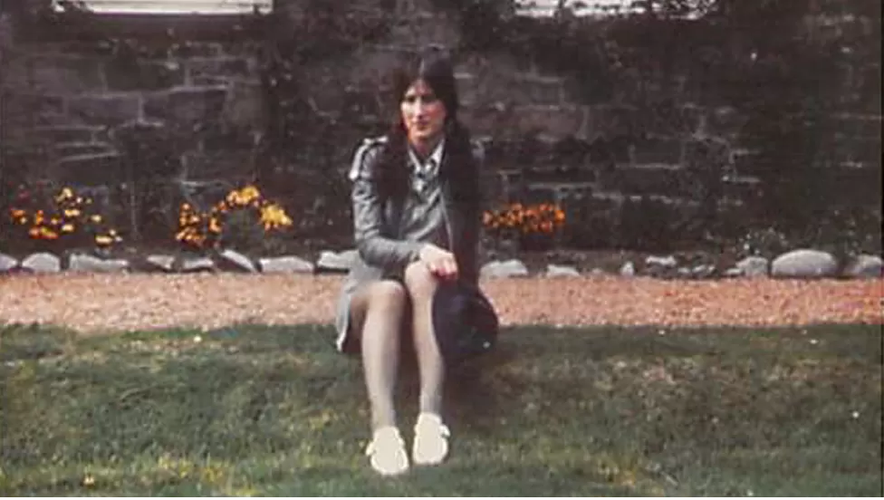 jovem branca de cabelo preto comprido sentada em frente a um muro