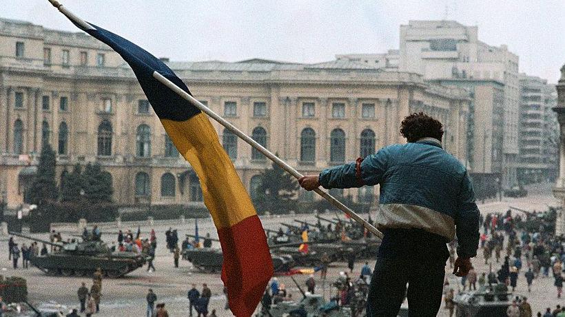 Hombre con una bandera de Rumania en la mano subido a un edificio, al fondo se ve una plaza llena de gente y de tanques. 