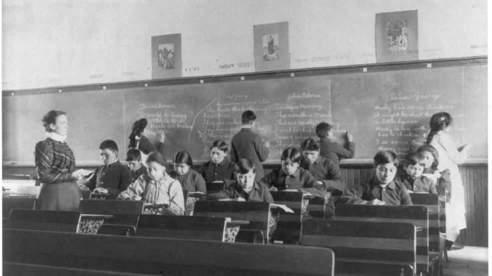 Crianças indígenas em sala de aula, algumas delas sentadas, e outras escrevendo na lousa