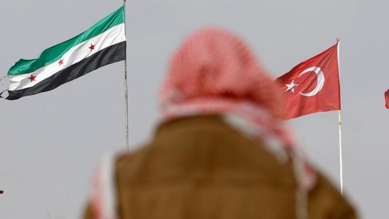 رجل يقف أمام العلم التركي وعلم الجمهورية السورية الأولى الذي يُمثل الثورة