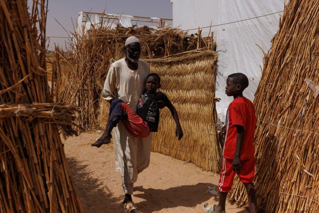 بخيت هارون يحمل ابنه الذي فقد القدرة على استخدام ساقيه بعد إصابته برصاصة في ظهره على يد قوات الدعم السريع العام الماضي في الجنينة بدارفور. الصورة في مخيم أمبيليا بالقرب من أدري، تشاد، بتاريخ 23 أبريل/نيسان 2024.