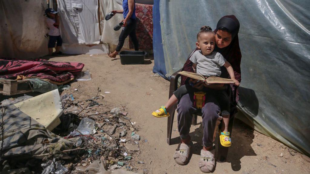 Mulher lê livro para criança no colo em meio a assentamento improvisado