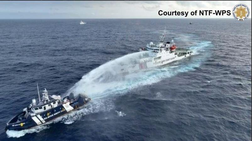 Um navio chinês azul marinho disparando um canhão de água em direção a um navio maior, branco e vermelho das Filipinas. Eles estão no mar, nenhuma terra é visível. Dois outros navios podem ser vistos à distância.