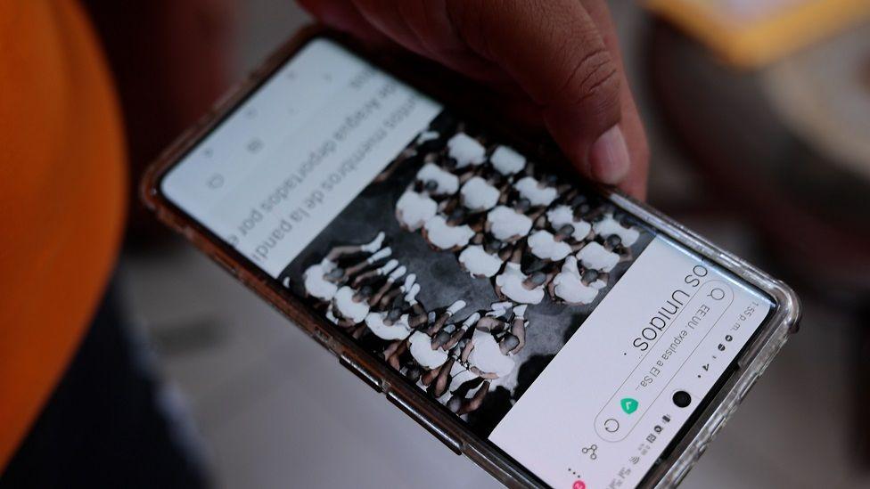 Un celular con una foto de un grupo de detenidos vestidos de blanco sentados en el suelo