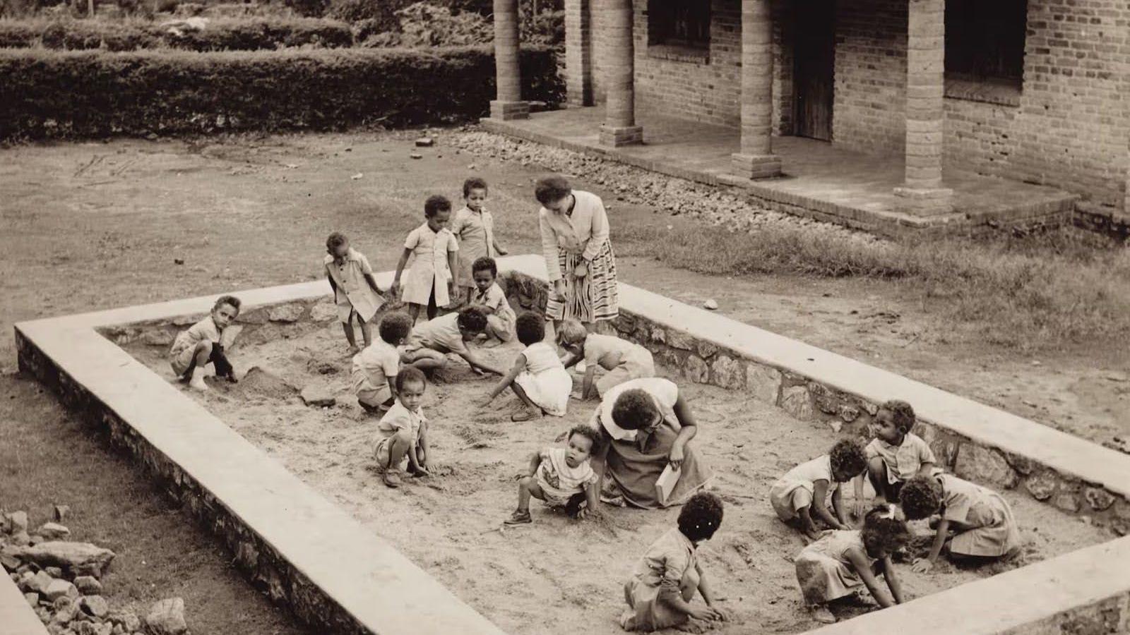 Un grupo numeroso de niños juega en un arenal. Hay dos mujeres con ellos.