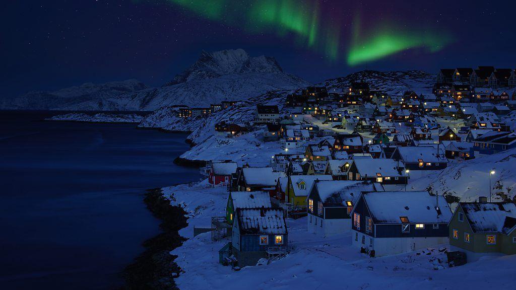 Casas cobertas de neve à beira mar e com uma aurora boreal verde no céu noturno acima delas