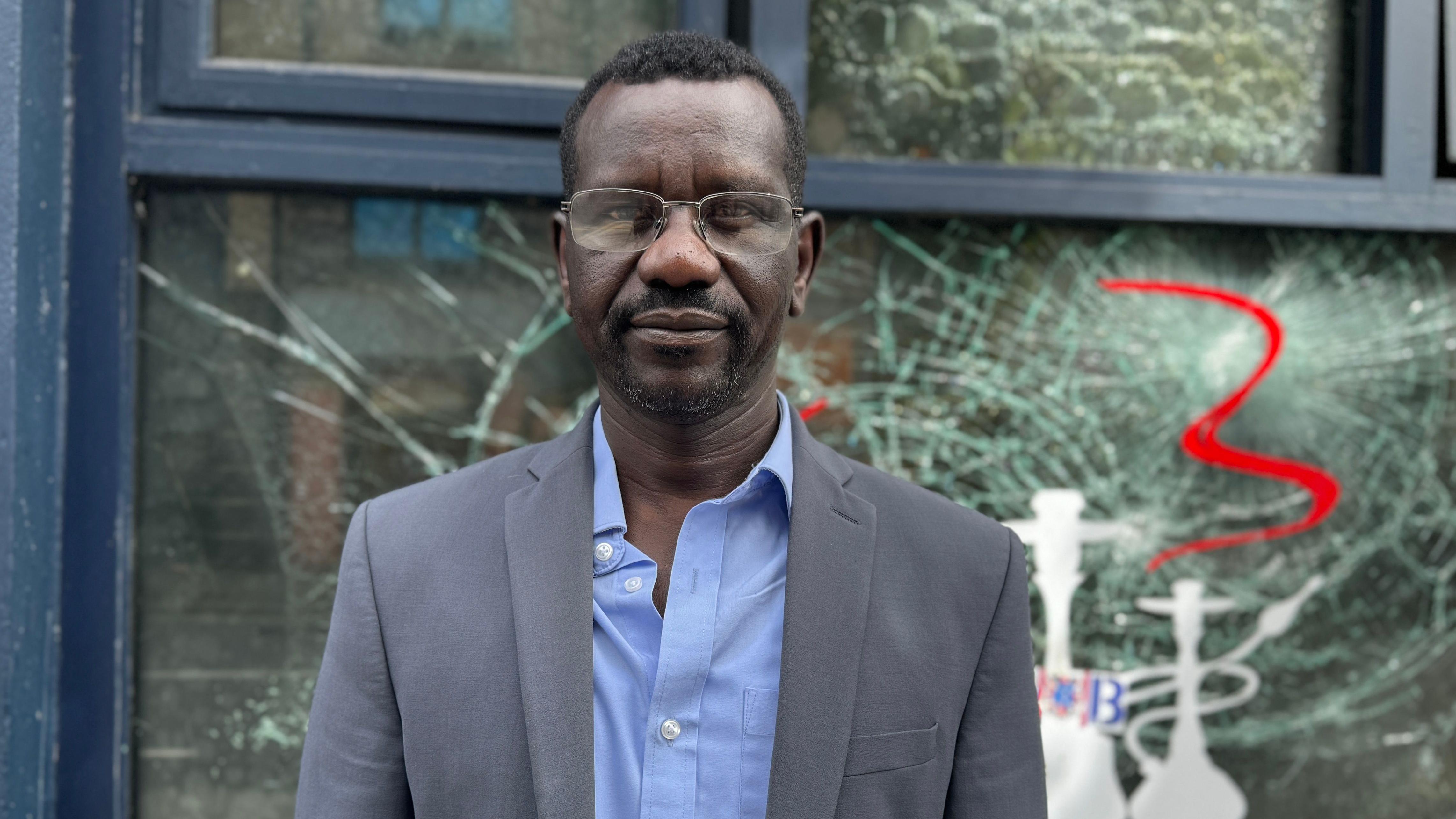 Mohammed Idris stands in front of his smashed shop window, wearing a blue shirt, grey suit jacket, and glasses.