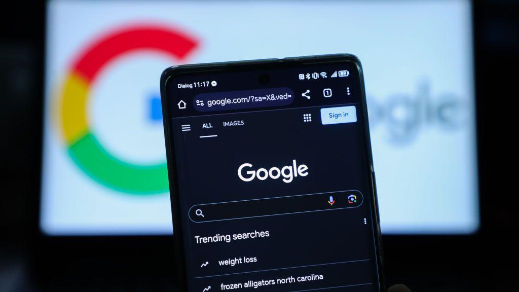 celular en el que aparece un buscador de Google. Atrás aparece el logo de la empresa en un computador