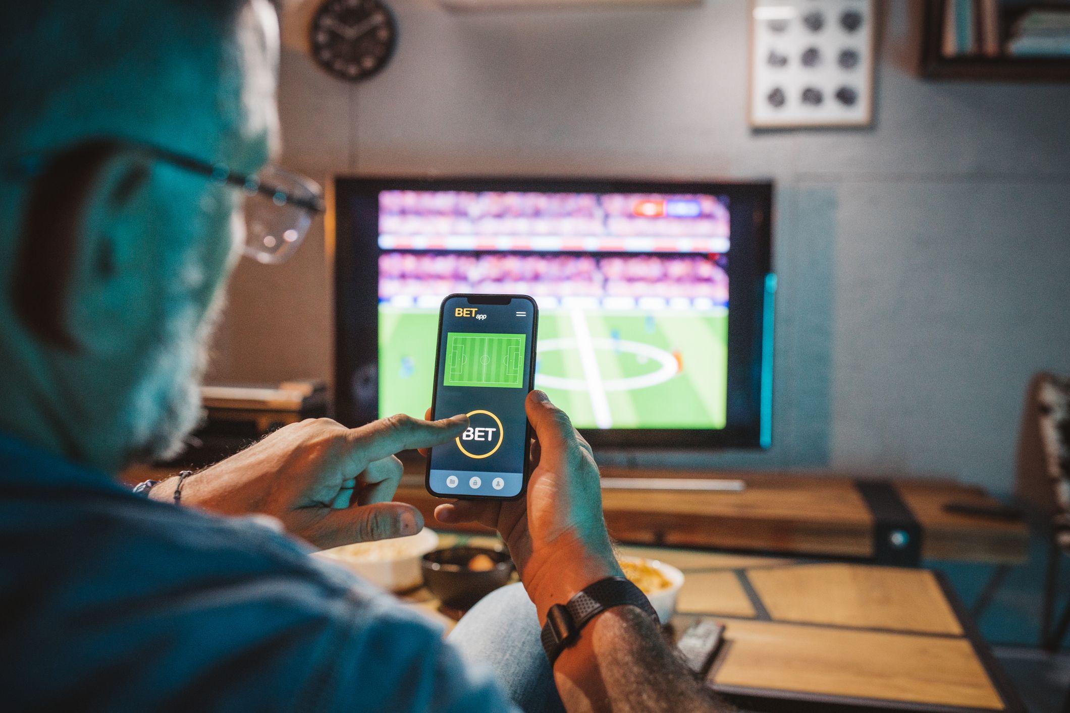 Fotografia colorida mostra um homem branco de óculos apostando em um jogo esportivo usando um aplicativo em seu celular, com uma televisão exibindo um jogo de futebol ao fundo