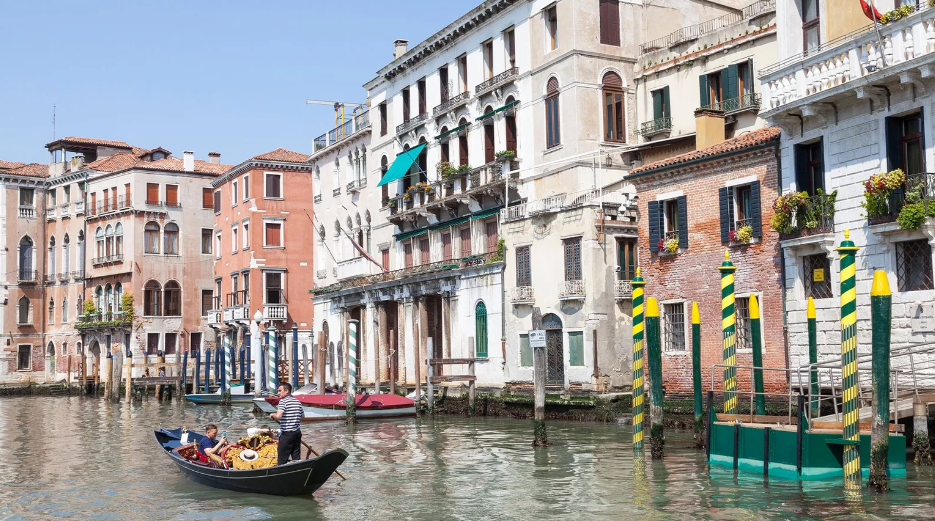 Sandolo navega em um canal com pessoas a bordo e casas ao fundo