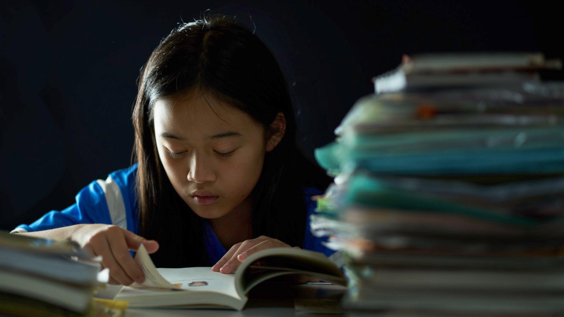 Menina asiática lendo um livro, com uma pilha de cadernos sobre a mesa