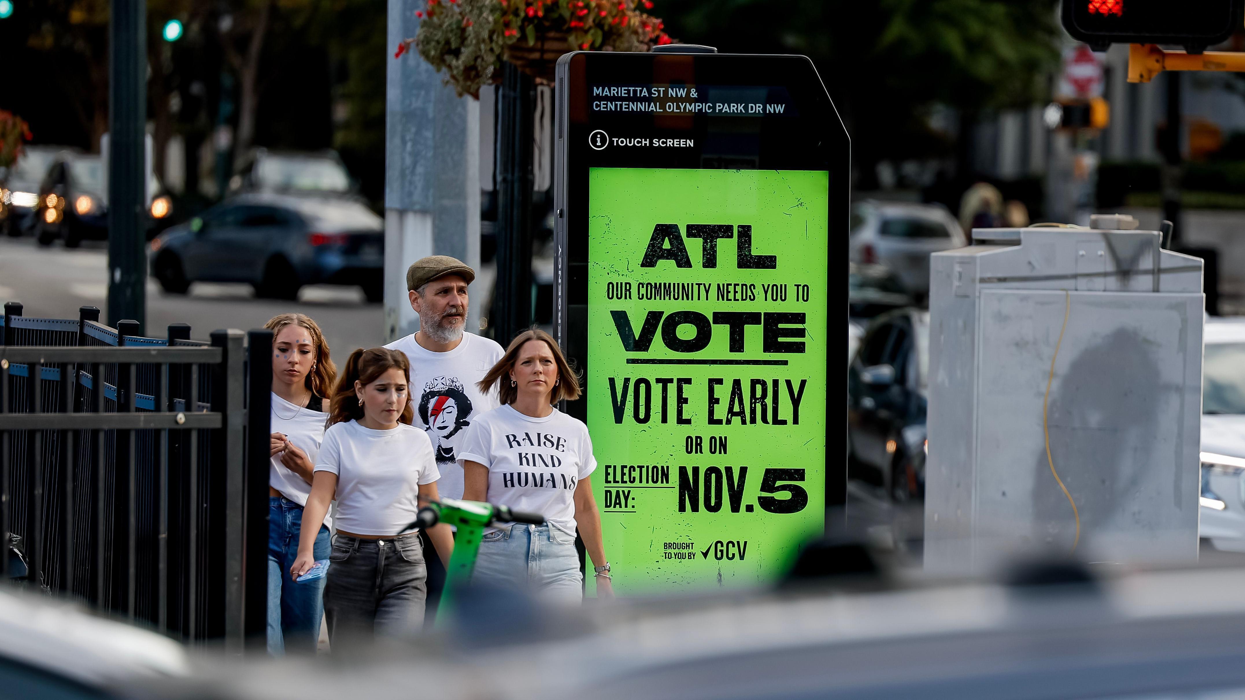 A foto mostra quatro pessoas caminhando na rua e ao lado delas uma placa onde se lê: ATL (Atlanta) nossa comunidade precisa do seu voto. Vote cedo. Dia da eleição: 5 de novembro