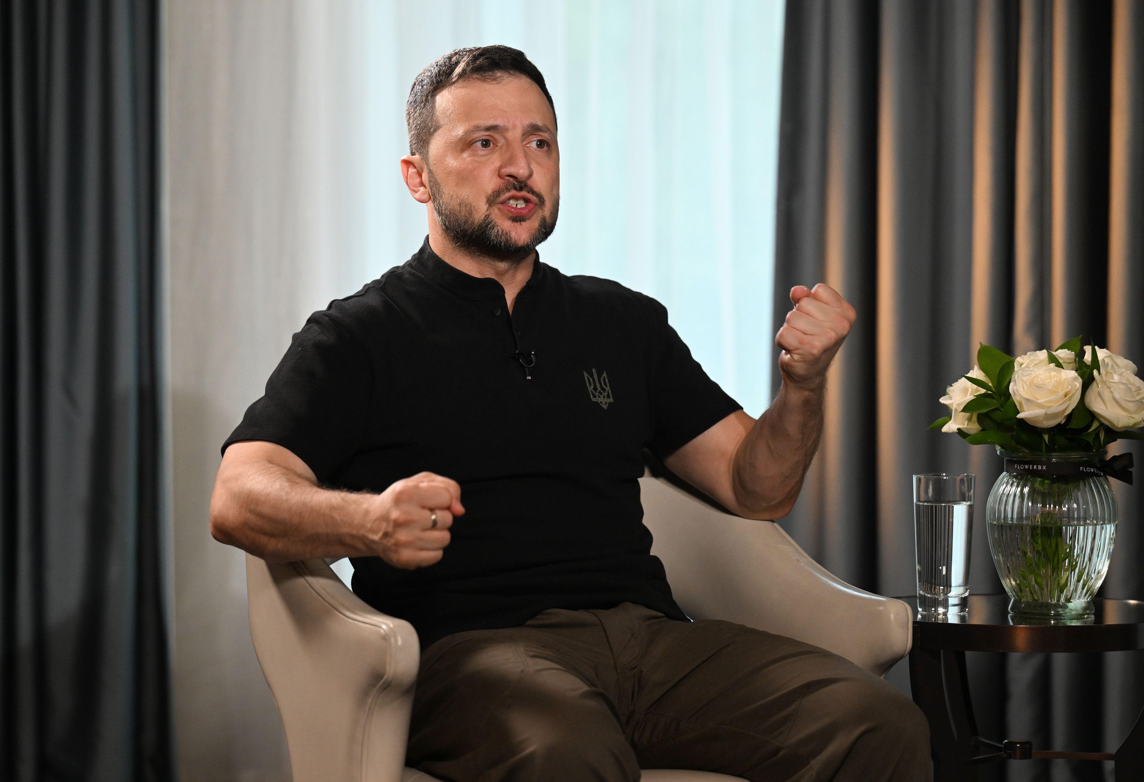 Volodymyr Zelensky é um homem de pele clara, de cabelos curtos escuros e barba cerrada. Ele está sentado em uma poltrona gesticulando. Está vestindo uma camiseta polo preta e uma calça escura. 