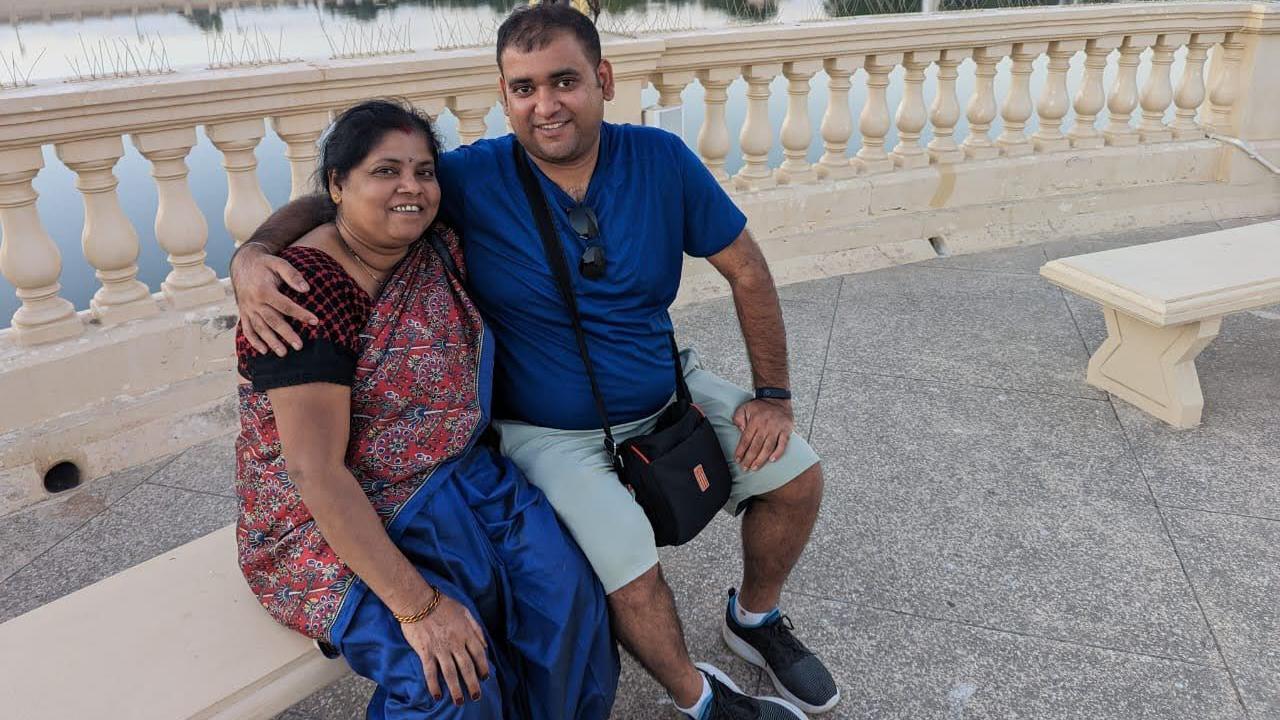 Atul Subhash com sua mãe em tempos mais felizes - ambos estão sentados em um banco de cor creme ao ar livre. Ele está vestindo uma camiseta azul, shorts, tem o que parece ser uma bolsa de câmera pendurada no pescoço e está sorrindo para a câmera com o braço em volta da mãe. A mãe dela está usando um vestido vermelho tradicional e também sorrindo para a câmera