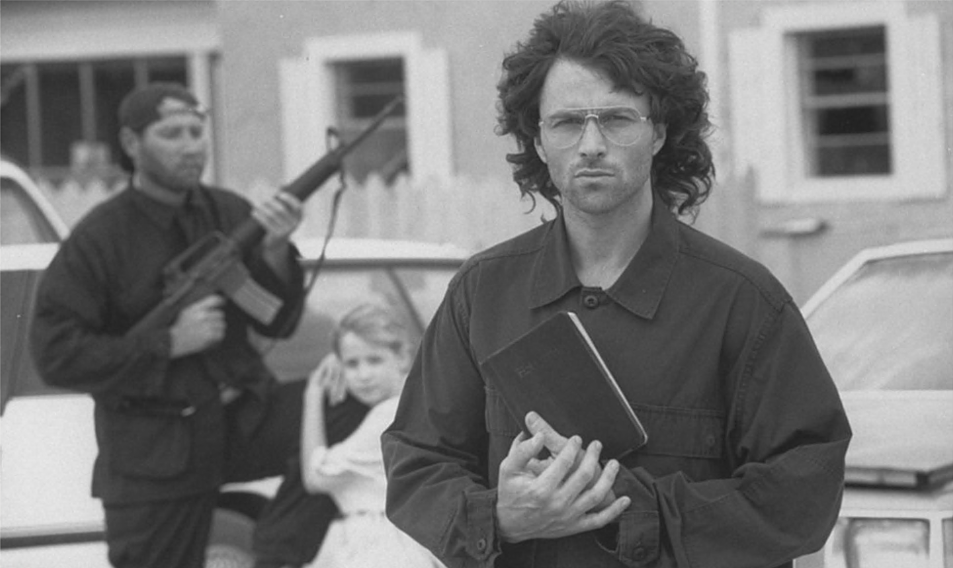 O ator Tim Daly no papel de David Koresh segurando uma bíblia em uma cena do filme 'Cerco ao Fanático do Texas', com um homem segurando uma arma e uma criança pequena ao fundo