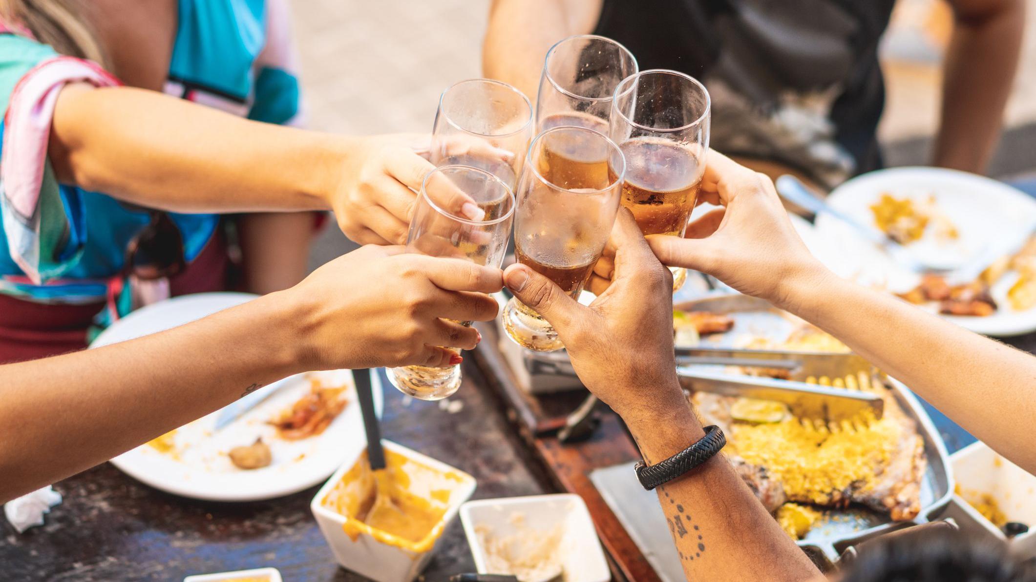 Mãos segurando copos de chopp e brindando, com uma mesa cheia de restos de comida ao fundo