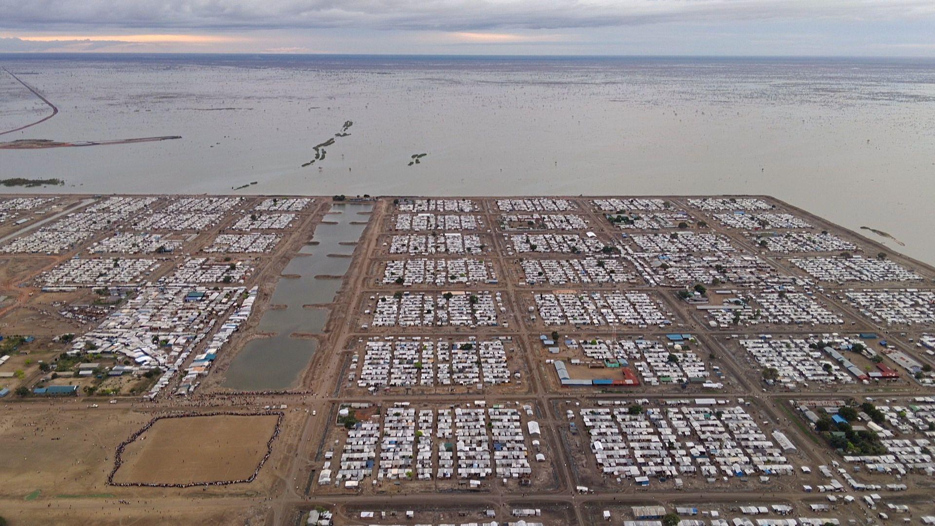 Imagem de drone de parte de um acampamento retangular cercado por água e coberto por cabanas apertadas
