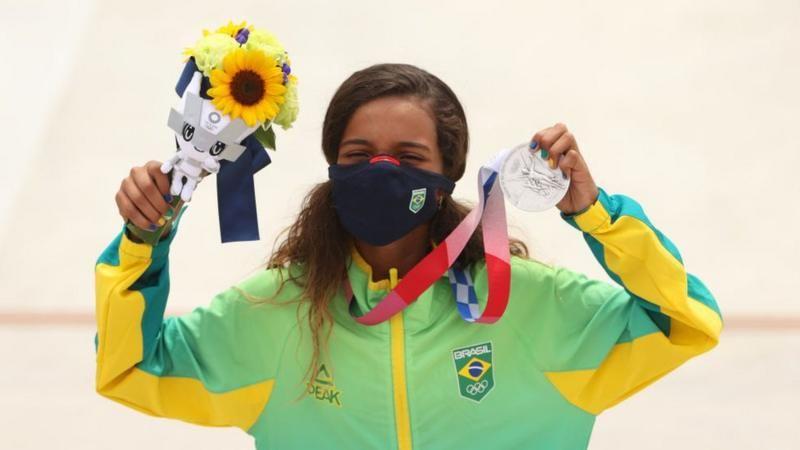 Rayssa Leal segurando sua medalha de prata nos Jogos Olimpicos de Tóquio 2021, ela usa máscara contra a covid-19, e segura na outra mão um pequeno buquê de flores, onde se destaca um girassol