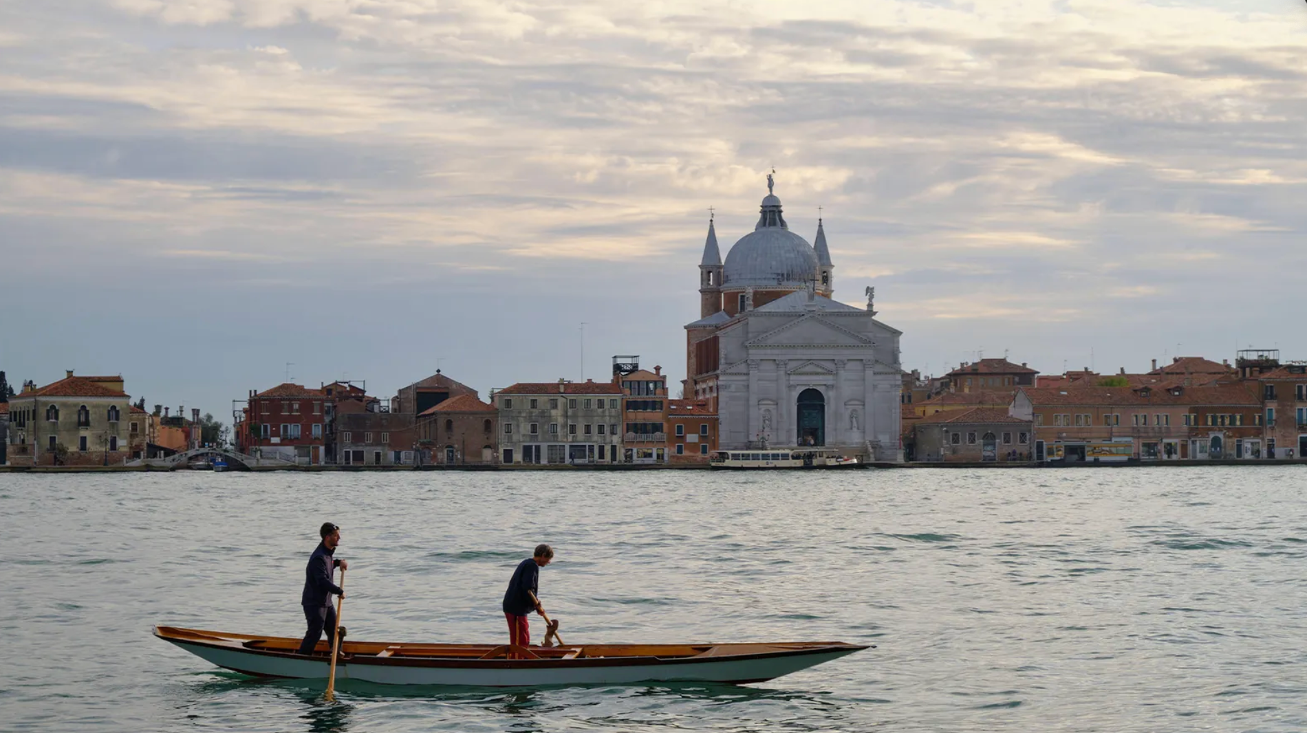 Dois homens em um barco navegam em Veneza, com a cidade ao fundo