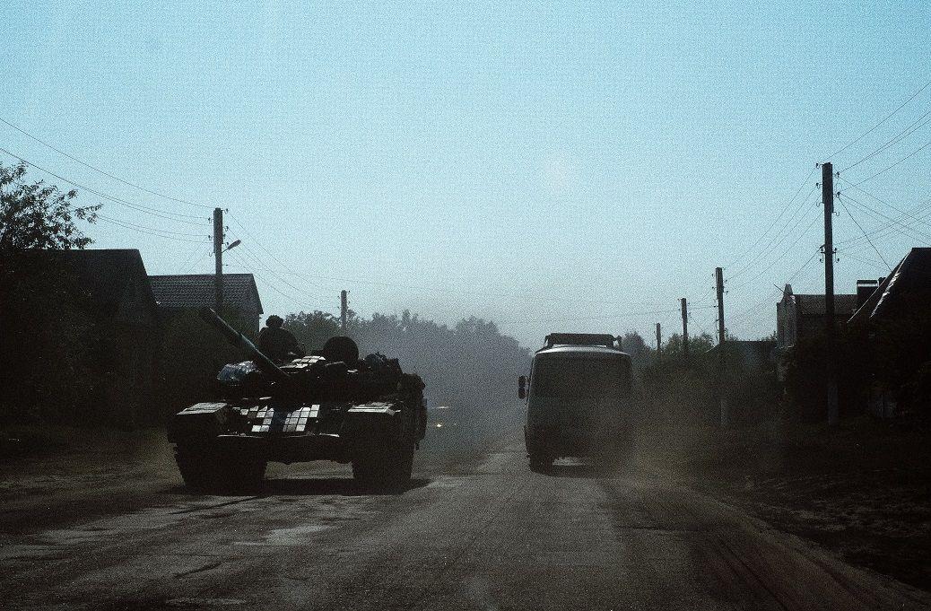 A foto mostra um tanque de guerra do lado esquerdo e um ônibus, do lado direito, em uma rua de terra. 