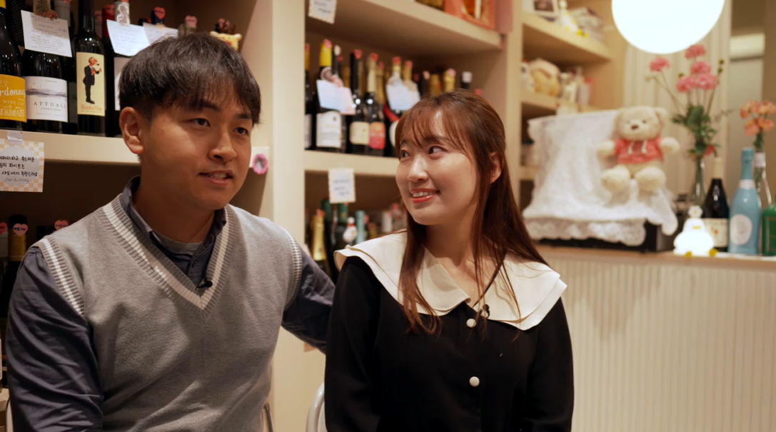 Tae Hyung e Min Jung sentados em uma loja de vinhos; ele fala e ela sorri ao olhar para ele 