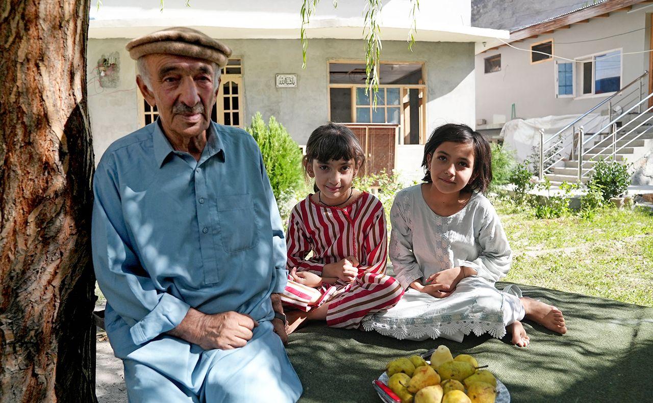 Sultan Ali, an elderly man, wears a cap and blue clothing while sitting next to his young granddaughters, who wear stripy and light blue clothing. They are sitting next to a plate of pears in a garden with a house in the background.