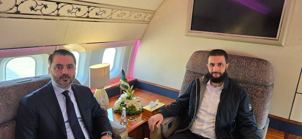 الشرع في الطائرة مرتدياً قميصاً أبيض وبنطالاً أسود دون ربطة عنق مع جاكيت غير رسمي، بجانبه الشيباني مرتدياً بدلة رسمية كحلية مع ربطة عنق بتدرجات اللون الكحلي، وفي المنتصف طاولة عليها العلم السعودي