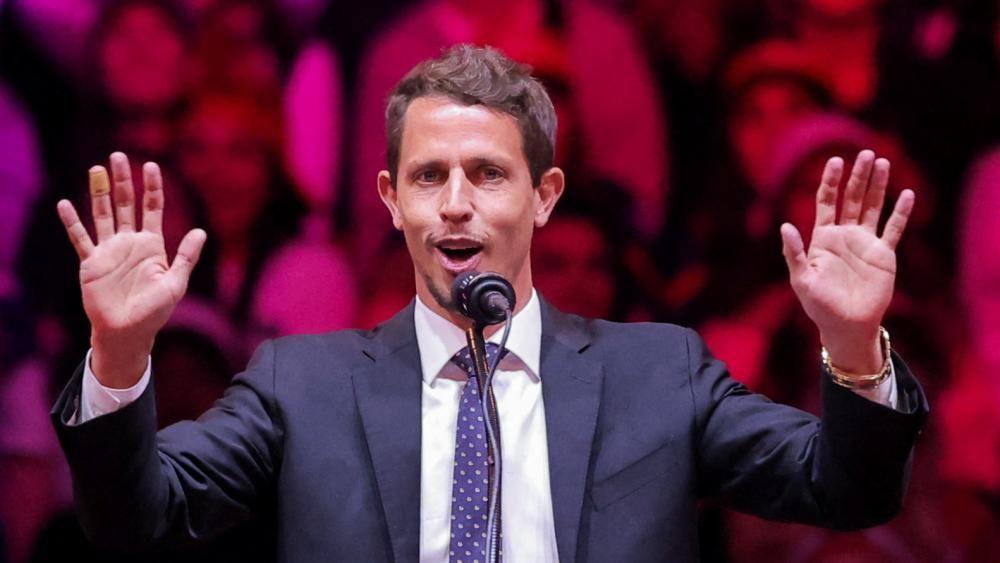 Foto mostra o comediante Tony Hinchcliffe em comício de Trump; ele está de terno, em um palco, com as mãos para cima enquanto fala em microfone