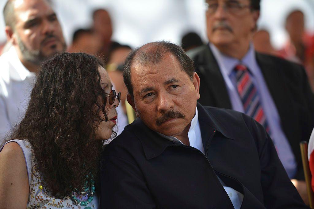 Daniel Ortega e sua esposa conversando em evento; eles estão sentados em uma plateia 