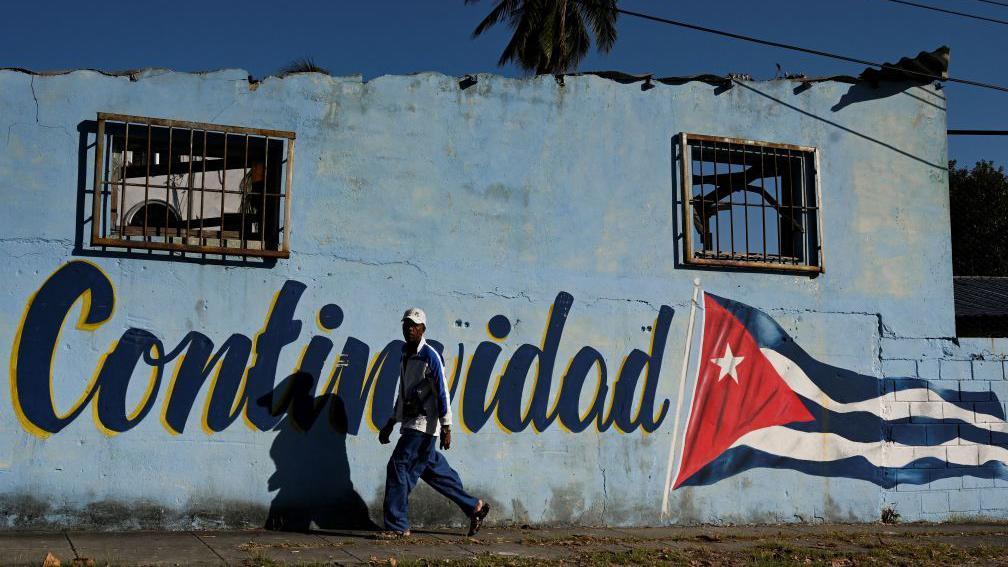 Um homem passa por uma parede pintada com a bandeira cubana que diz 