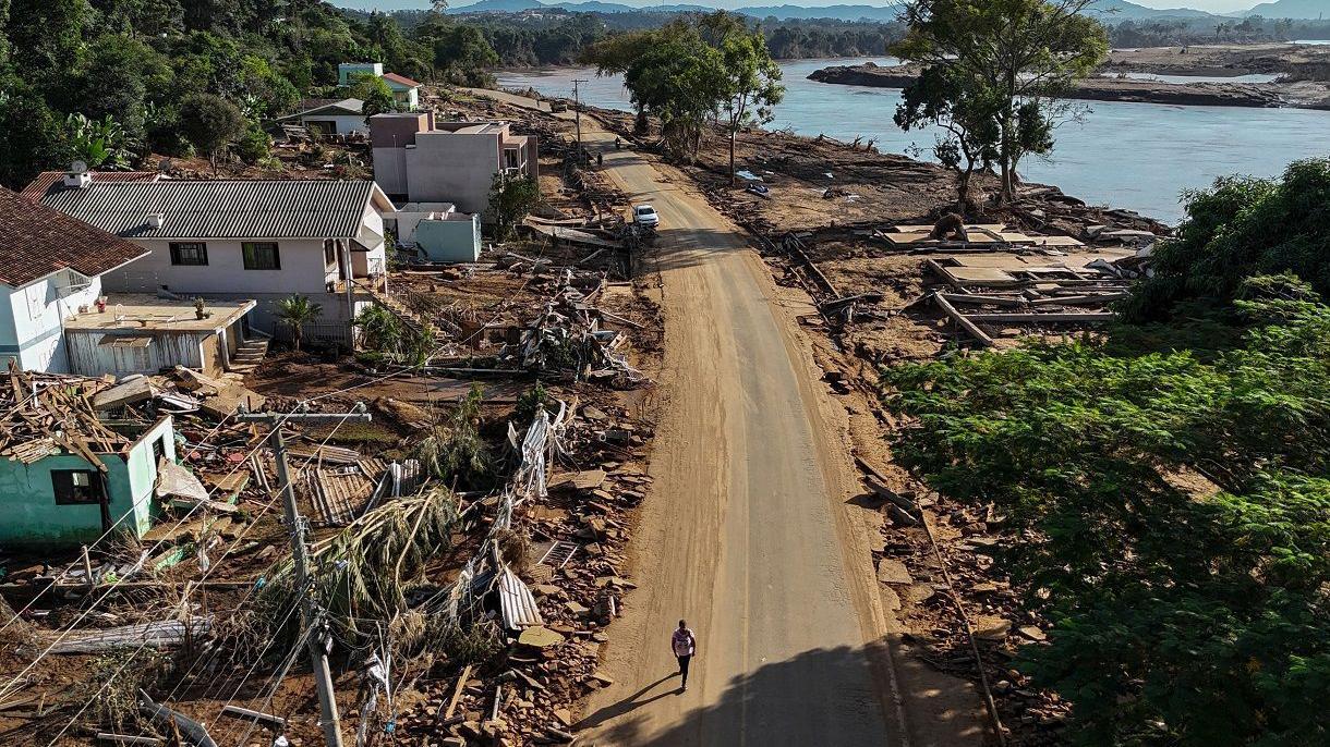 Foto aérea mostra pessoa andando em rua perto de casas destruídas; na margem da foto, está um rio